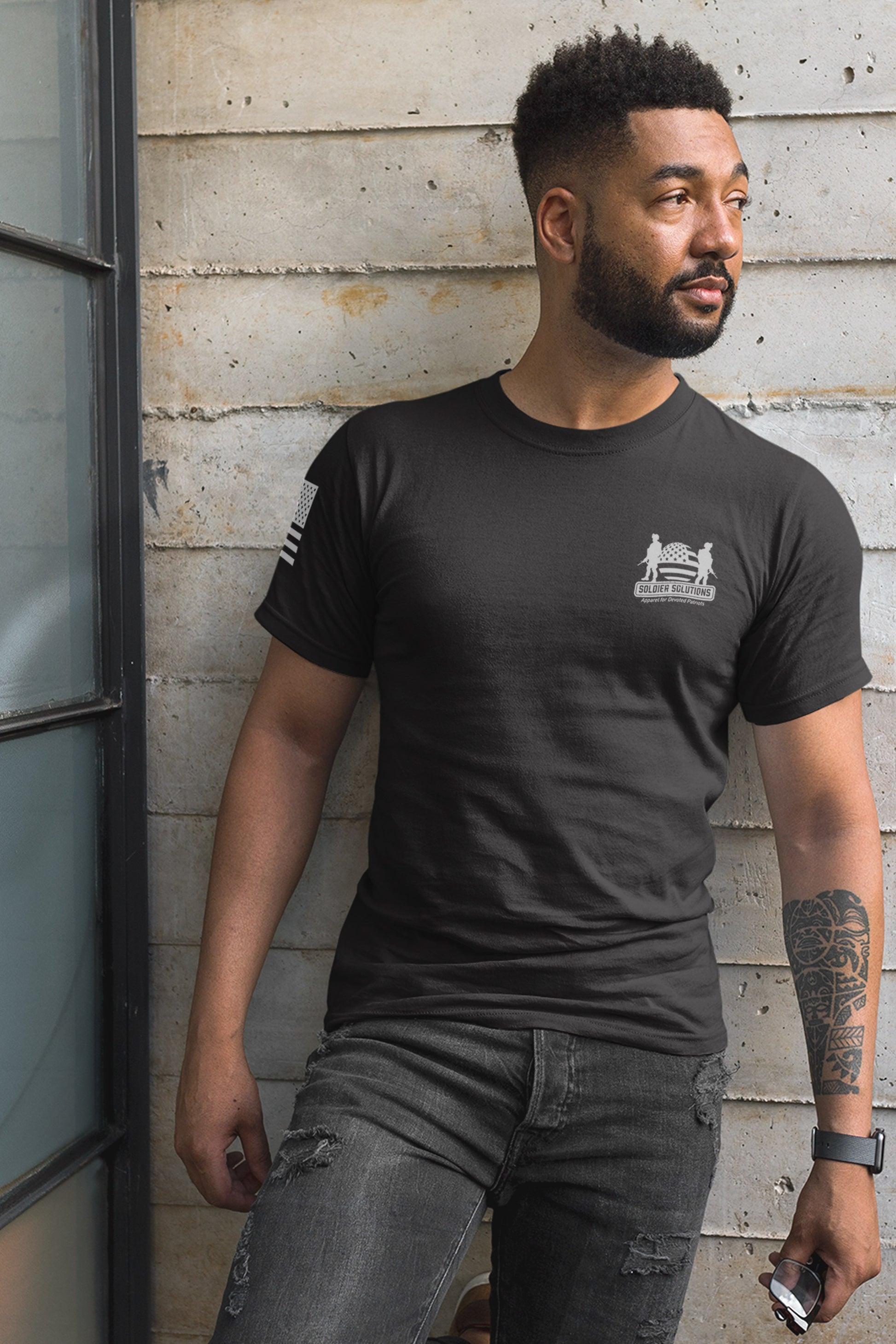 Man wearing black United We Stand 2nd amendment t-shirt by Soldier Solutions, featuring small chest logo and right sleeve flag print, standing casually in front of industrial wall.
