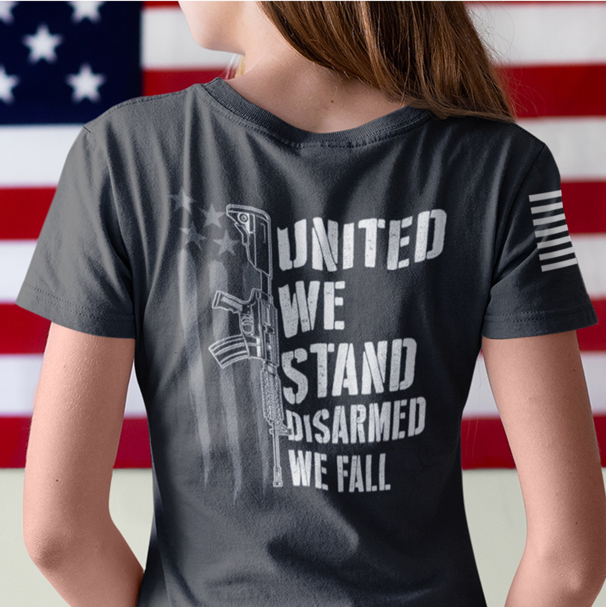 Close up of the back of woman wearing patriotic 2nd Amendment shirt with white rifle graphic and bold text ‘United We Stand, Disarmed We Fall’ over distressed American flag.