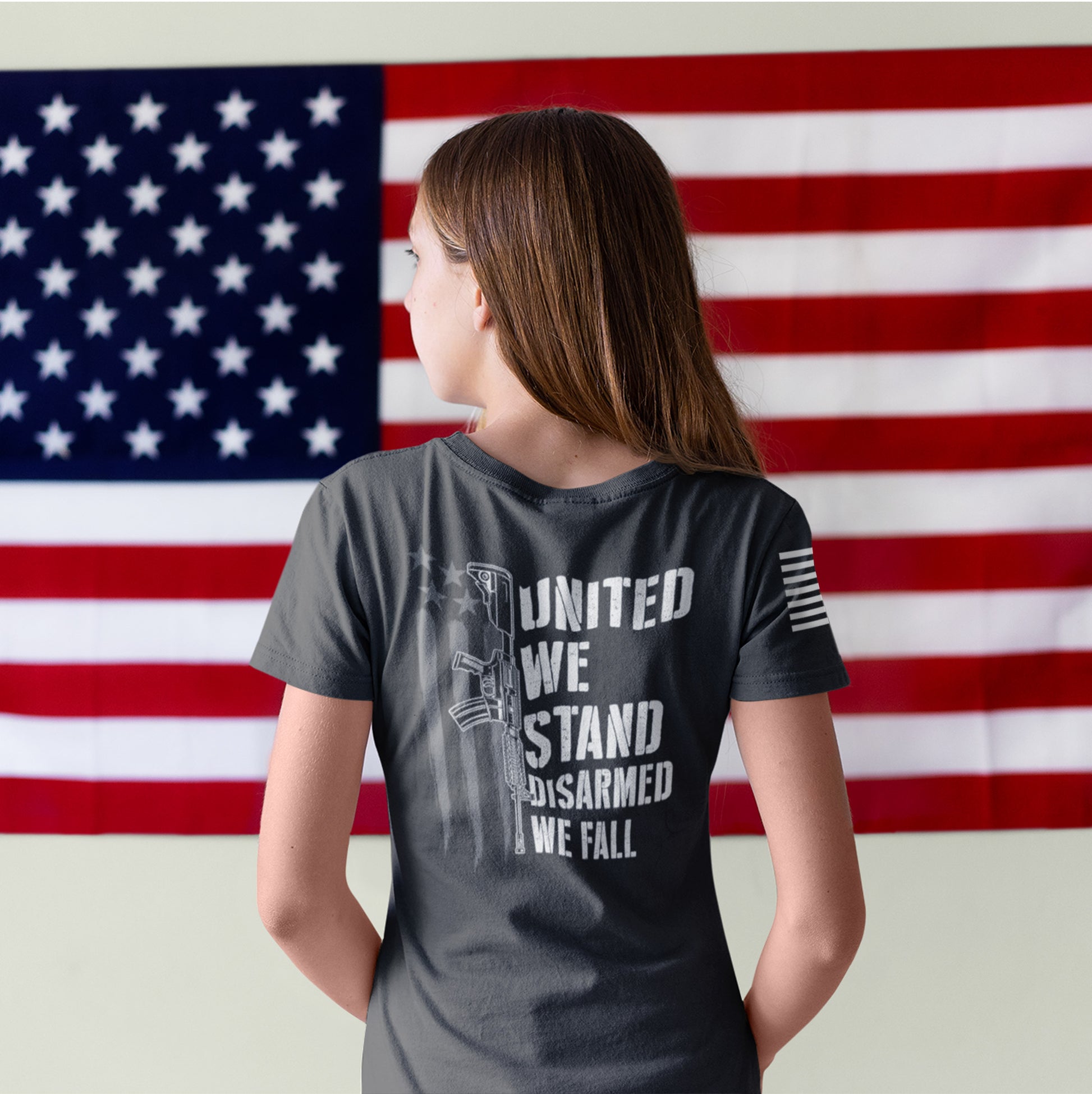 Back of woman wearing patriotic 2nd Amendment shirt with white rifle graphic and bold text ‘United We Stand, Disarmed We Fall’ over distressed American flag.