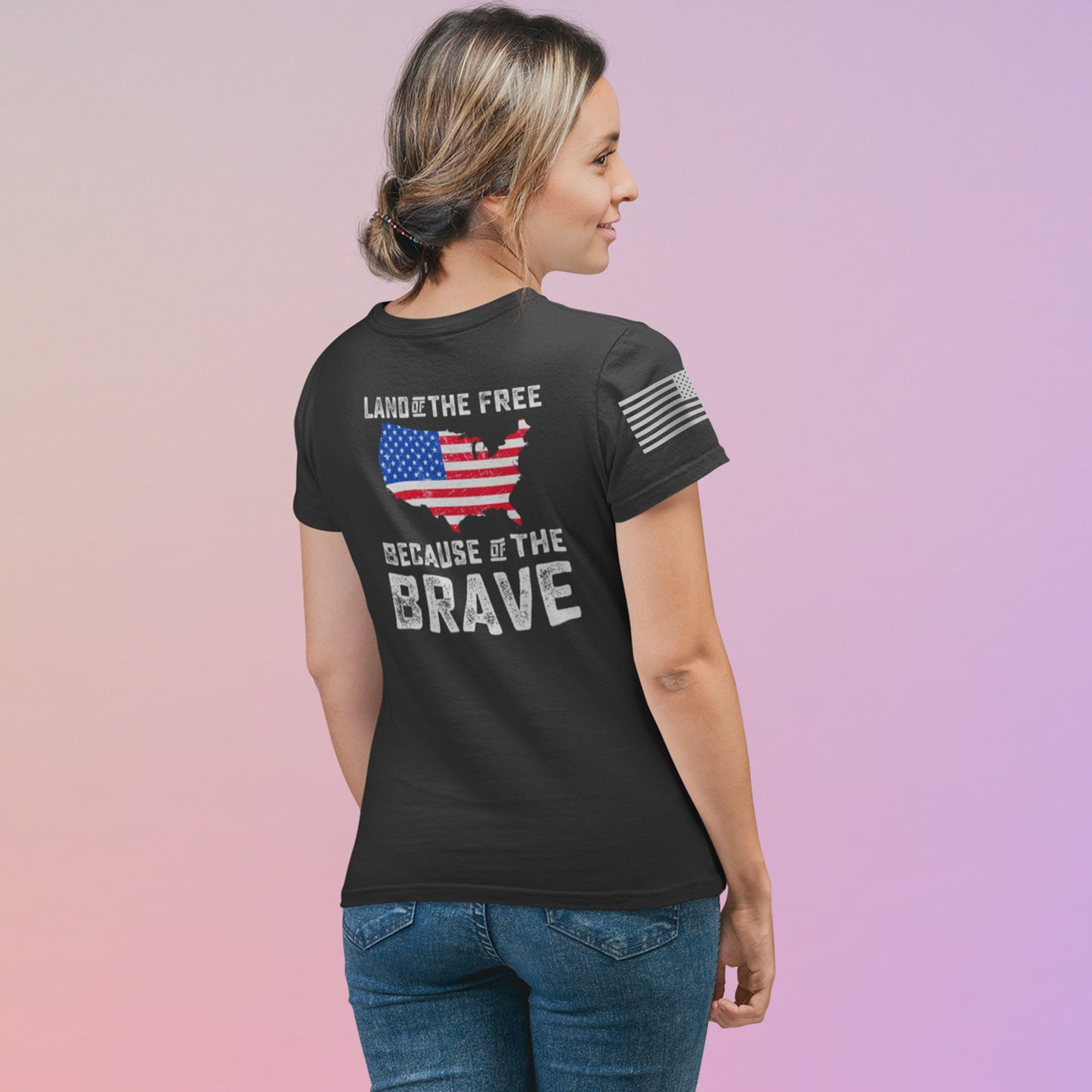 Back view of woman wearing Land of the Free Because of the Brave t-shirt with U.S. map graphic in American flag colors and distressed bold text.
