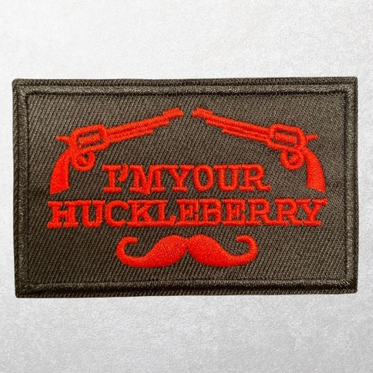 Hook-and-loop tactical patch with “I’m Your Huckleberry” text, crossed revolvers, and mustache embroidery. Black and Red