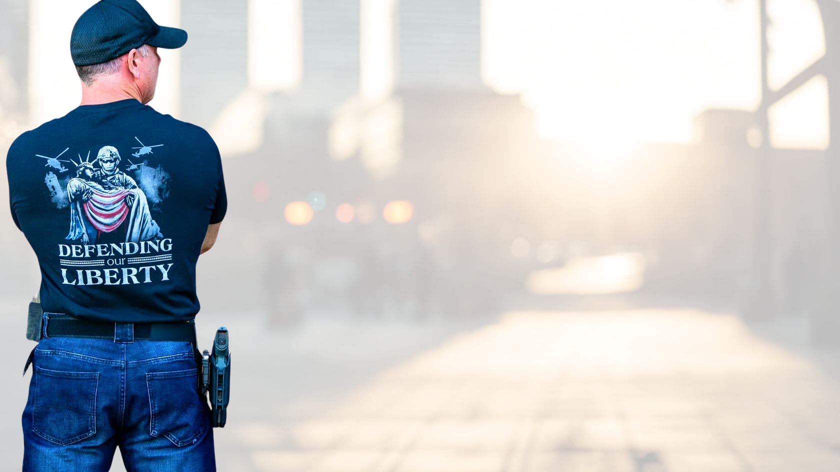 Veteran wearing a Soldier Solutions Defending Our Liberty t-shirt with a patriotic graphic, standing confidently in a city street at sunrise.
