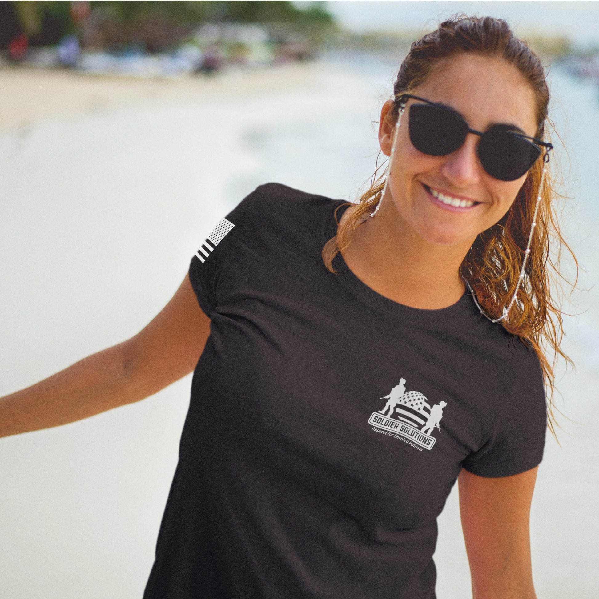 Smiling woman wearing Let Freedom Ring t-shirt by Soldier Solutions, front view with Soldier Solutions chest logo and sleeve flag graphic, walking on the beach.